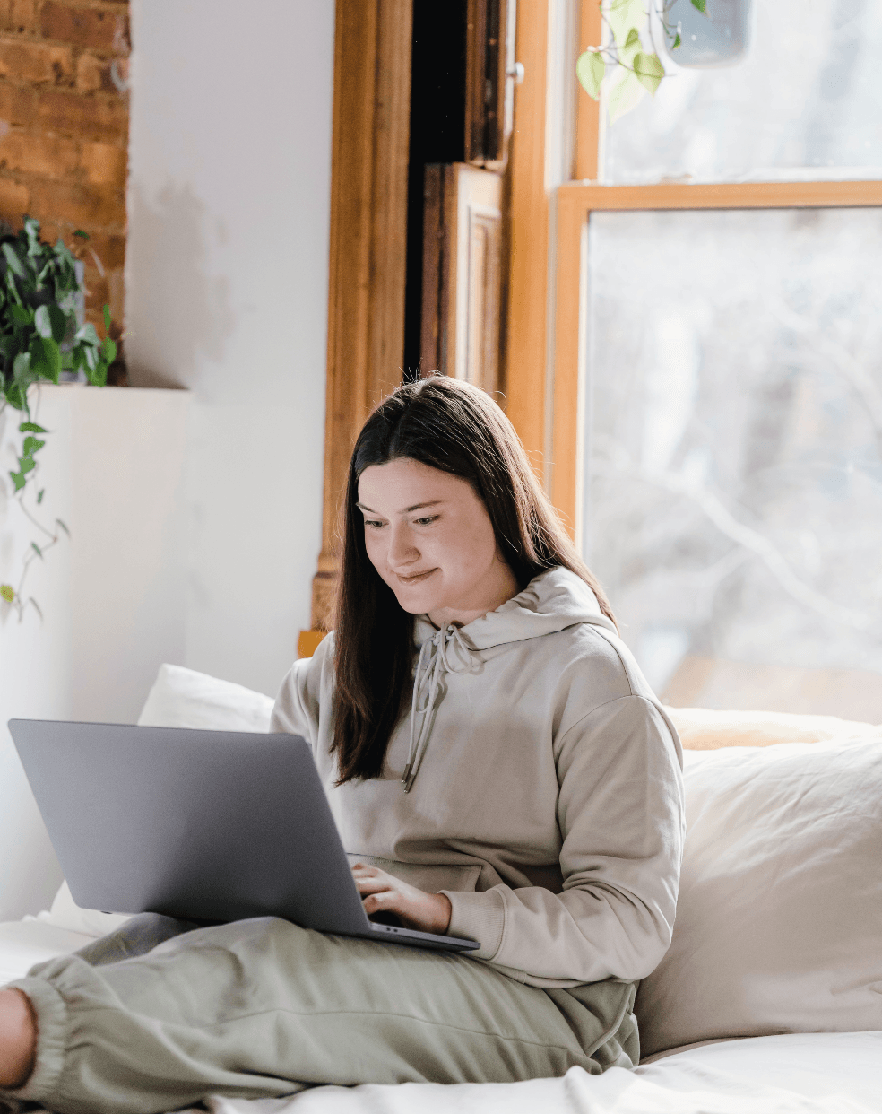 girl working on a laptop while seated on a bed in a sunny room